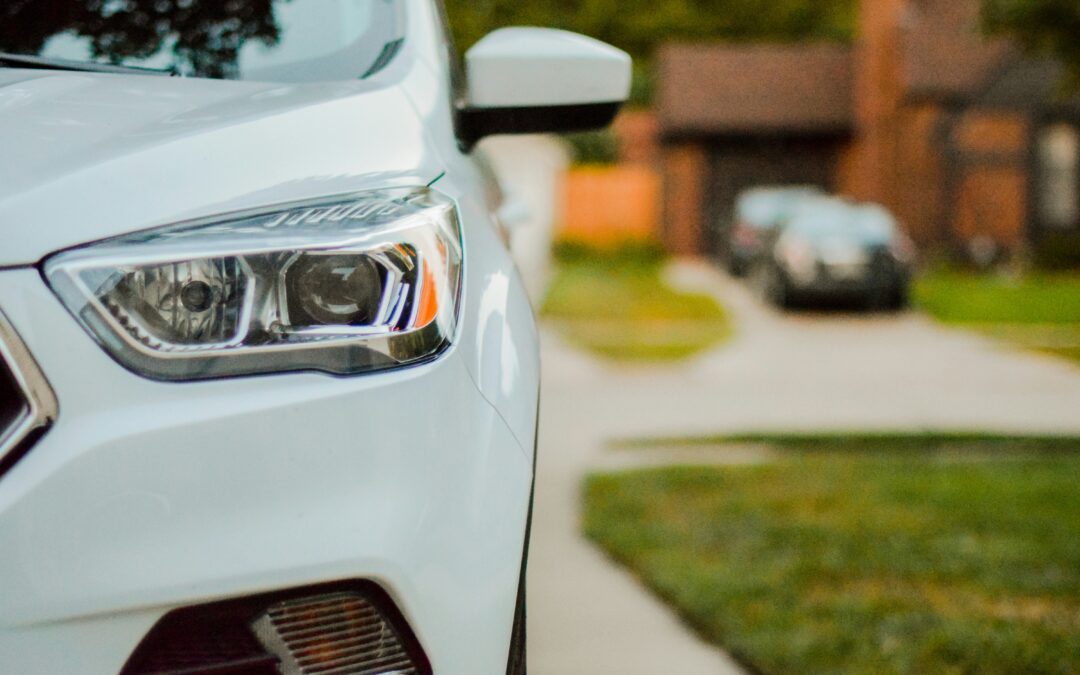 Close-up of the front headlight and fender of a white car parked in a driveway, with a blurry suburban house and another car in the background.